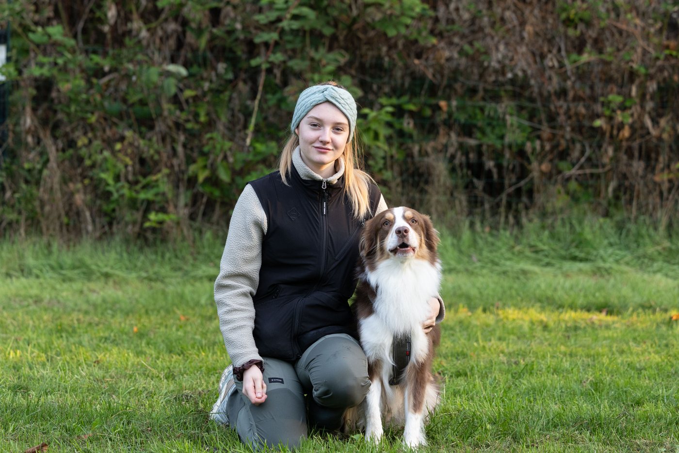 Johanna kniet auf der Wiese neben ihrem Australian Shepherd Cooper